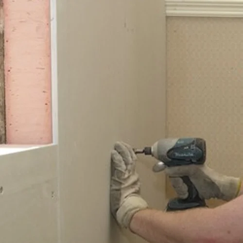 Worker using a power drill to install an insulated plasterboard panel.